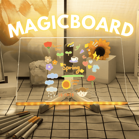 A MagicBoard product with a transparent surface displaying colorful drawings including a sun, flowers, and the words 'spring'. Below the board, there are markers and a microfiber cloth.
